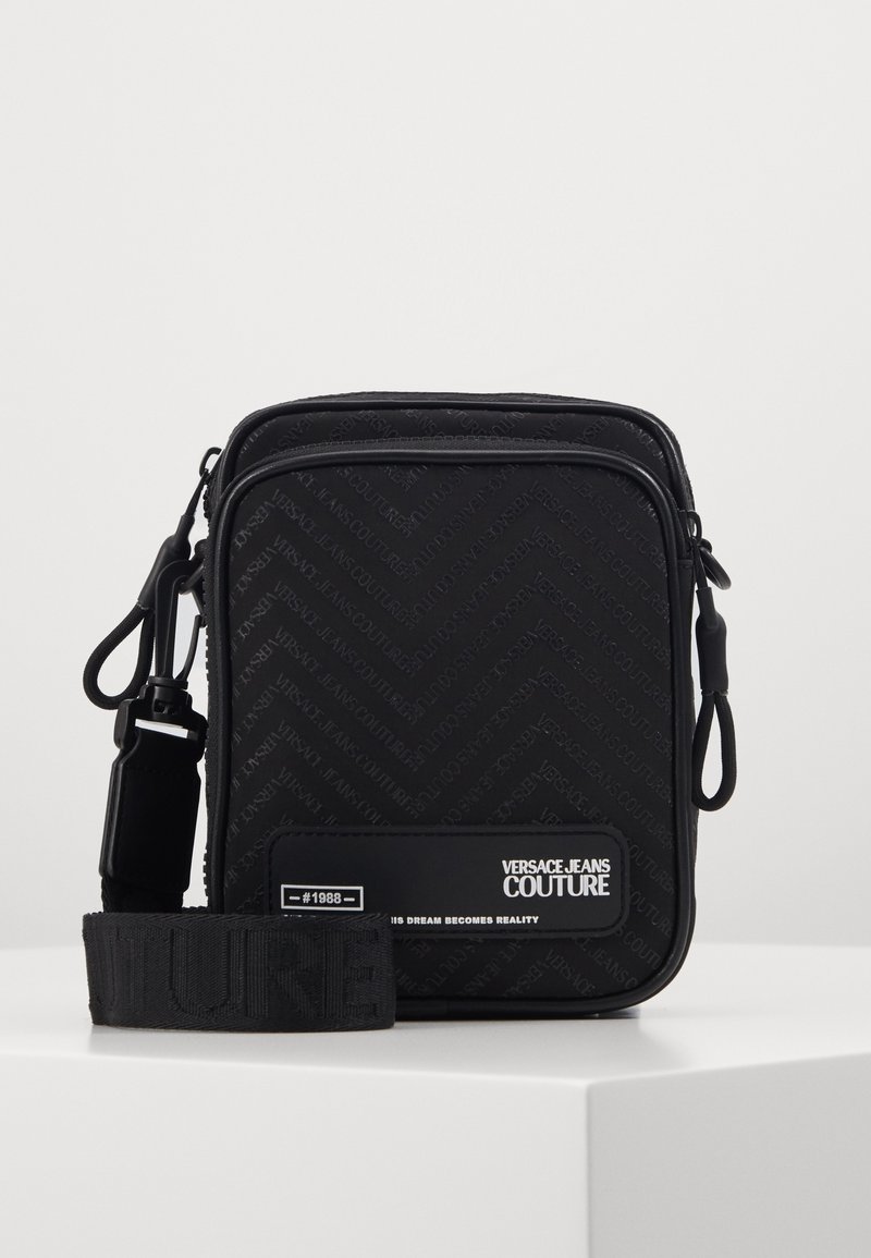 Black crossbody bag with a chevron pattern, zip closure, and a detachable strap featuring textured branding accents.