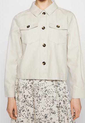 Veste beige clair boutonnée avec deux poches poitrine portée sur une jupe blanche à imprimé floral noir.