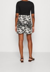 Musta istuva yläosa yhdistetty vaaleanruskeisiin shortseihin, joissa musta lehtikuvio. Sandaaleissa on matala lohkokorko ja ruskea yksityiskohta.