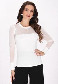 Maglietta bianca a maniche lunghe con scollatura e maniche in rete trasparente, caratterizzata da piccole perline e un dettaglio arricciato sul petto. Abbinata a pantaloni neri.