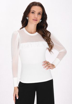 Maglietta bianca a maniche lunghe con scollatura e maniche in rete trasparente, caratterizzata da piccole perline e un dettaglio arricciato sul petto. Abbinata a pantaloni neri.