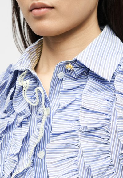 Κοντινό πλάνο μιας γυναίκας που φορά μια ριγωτή μπλούζα με βολάν σε μπλε και άσπρο, με ένα διακοσμητικό μαργαριταρένιο και χρυσό καρφί στο γιακά.