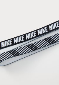 Juodas ir baltas elastingas kaspinas, turintis pakartotinį "Nike" tekstą vienoje pusėje ir dryžiuotą raštą kitoje pusėje, su minkšta tekstūra.