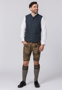 Traditionelles bayerisches Outfit mit einer strukturierten dunkelblauen Weste, braunen Lederhosen mit Stickerei und gestrickten grauen Kniestrümpfen.