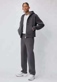 Sweat à capuche gris et pantalon de survêtement assorti, confectionnés en tissu doux. Dotés d'une fermeture éclair, de poches avant et d'une coupe décontractée. Portés avec des baskets blanches.