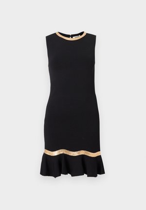 Nő áll, fekete ujjatlan ruhát visel arany díszítéssel a nyakkivágásnál és a fodros szegély felett, fekete loaferekkel párosítva.