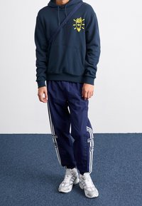 Laudan sininen huppari, jossa keltainen grafiikka ja kengurulompakko; laudan siniset treenihousut, joissa valkoiset raidat ja logot; harmaat lenkkarit. Kankaan pinta on teksturoitu.