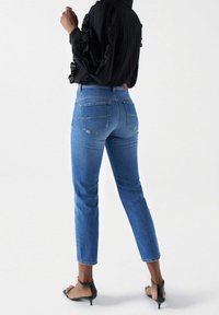 Jeans de mezclilla azul de talle alto con corte ajustado, ligero desgaste y bolsillos traseros, combinados con una blusa negra texturizada y calzado con tiras.