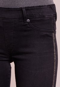 Los vaqueros de mezclilla negros presentan un corte ajustado, con un bolsillo lateral con cremallera adornado con pequeños acentos metálicos. Textura suave, diseño clásico de cinco bolsillos.