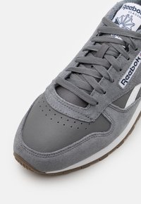 Zapatilla gris que presenta una mezcla de ante y cuero, puntera perforada, cordones planos y un detalle en blanco en la lengua.