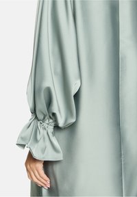 Blouse en satin vert atténué avec de longues manches bouffantes et des poignets froncés. Texture lisse avec un léger brillant, présentant une silhouette décontractée.