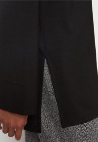 Schwarzes Langarmshirt mit seitlichem Schlitz, kombiniert mit grauem Hahnentritt-Muster. Glatte Textur, lässiges Design und lockere Passform.