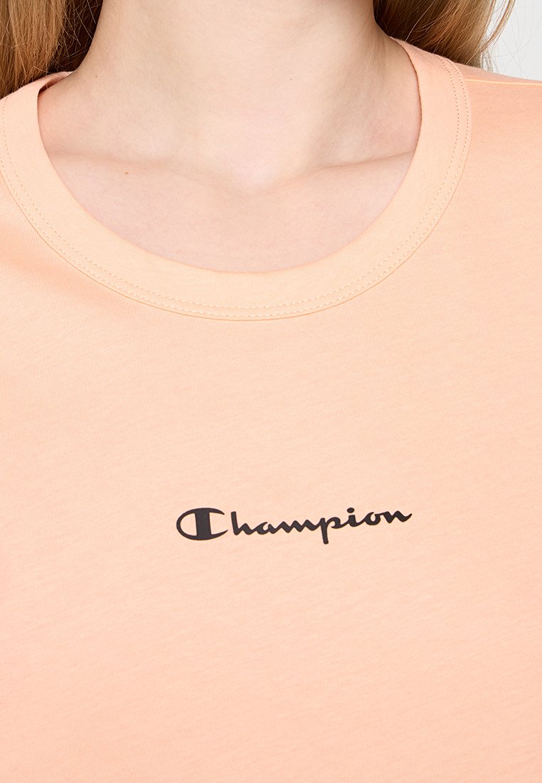 T-shirt in cotone pesca con collo rotondo. Presenta un logo "Champion" nero sul davanti. Texture liscia con una vestibilità rilassata.
