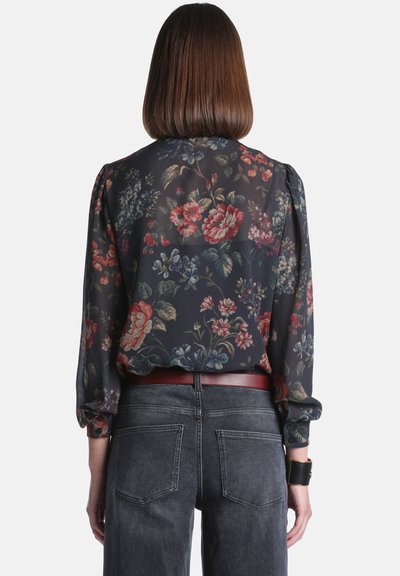 Femme aux cheveux bruns raides portant un chemisier noir transparent à fleurs rentré dans un jean gris foncé avec une ceinture marron et un large bracelet noir.