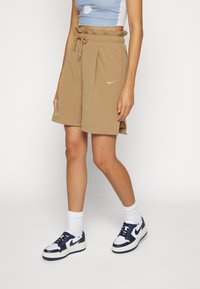 Světlé, volně střižené šortky v tan barvě s elastickým pasem a šnůrkou, sešívané s malým bílým logem Nike. Doplňují je tmavé tenisky a bílé ponožky.