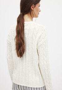 Weißer Strickpullover mit vertikalen Rippen, langen Ärmeln und geradem Saum. Die Textur ist weich und dick, geeignet zum Schichten.