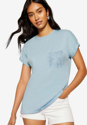 T-shirt en coton bleu clair avec des manches retroussées et une poche en crochet texturée, associé à un short blanc. Tissu lisse et coupe décontractée.