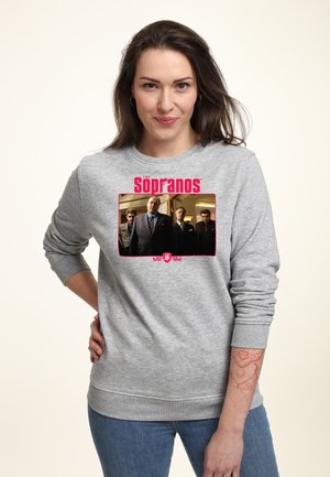 Femme aux longs cheveux bruns portant un sweat gris affichant une photo et le logo du casting de The Sopranos, debout, la main sur la hanche, sur un fond clair.