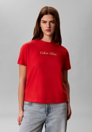 Kvinde iført en rød Calvin Klein T-shirt med gyldent logo og lyseblå jeans, stående foran en ensfarvet grå baggrund.