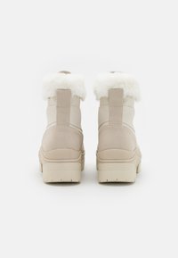 Botas de tobillo beige con un tejido texturizado, cuello de pelo sintético y una suela de goma gruesa. Cuenta con una pestaña en la parte posterior para facilitar el uso.