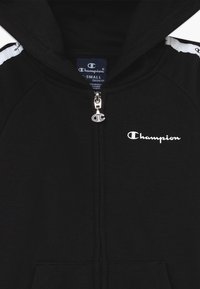 Felpa nera con zip Champion, logo bianco sul petto, tirante della zip con emblemma "C" e finiture bianche sulle spalle.