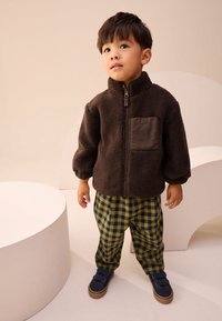 Veste polaire marron avec une poche poitrine, pantalon à carreaux verts et noirs, et baskets bleues avec des bandes auto-agrippantes sur un fond clair.