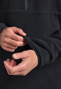 Felpa nera in pile con scollo a mezza zip, dotata di polsini a coste. Il tessuto strutturato è morbido e caldo, mentre si vedono mani che regolano le maniche.