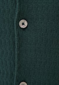 Vihreä neulottu kangas, jossa on teksturoitu kuvio, ja jossa on pyöreä, kirkas nappi, jossa on neljä reikää. Lähikuvassa korostuvat yksityiskohdat.