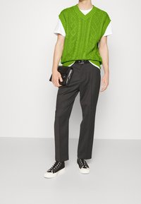 Gilet en tricot vert porté sur un t-shirt blanc, assorti à un pantalon sombre ajusté et des baskets noires. Tenant une pochette noire.