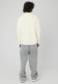 Un pull crème avec un col et des poignets côtes, porté sur une chemise blanche, associé à un jean gris ample et des baskets blanches.