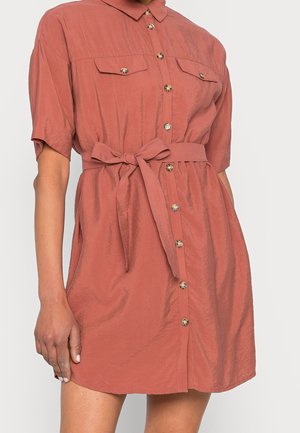 Šaty košilového střihu s krátkými rukávy v barvě rzi, s knoflíky vpředu, náprsními kapsami s klopami a zavazovacím páskem v pase na stojící osobě.