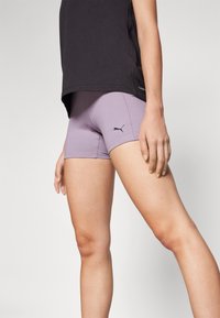Purpurowe szorty sportowe wykonane z elastycznego materiału, z czarnym logo Pumy z boku. Noszone z luźną czarną bluzą, ukazującą dopasowany krój.