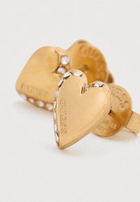 Σκουλαρίκια σε σχήμα καρδιάς από χρυσό με διακριτικά στρας και ανάγλυφο λογότυπο "GUESS". Λεία υφή, απλός σχεδιασμός.