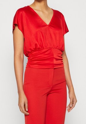 Femme portant un chemisier rouge vif à col en V et manches courtes avec une taille froncée, associé à un pantalon rouge taille haute.