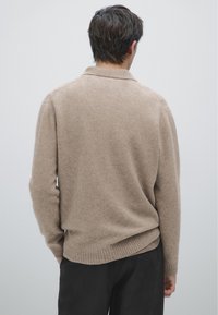 Beige tröja med texturerad stickning, med krage och ribbat avslut. Bärs med mörka byxor som visar en avslappnad passform och rena linjer.