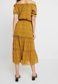 Senapsgul av-axeln klänning med puffärmar, blommönster, lagerdesign och flytande fåll, bärs med svarta klacksandaler.