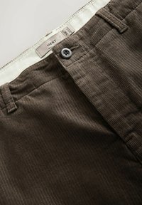 Nahaufnahme von braunen Cordhosen mit Knopfverschluss und sichtbarem inneren Taillenbandlabel, das die Größe 38R und die Marke NEXT zeigt.