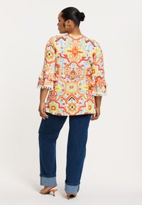 Felgekleurde top met uitlopende mouwen in oranje, geel en blauw, gecombineerd met donkerblauwe gerolde jeans en lichtoranje hakken.