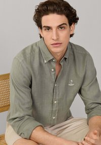 Jeune homme aux cheveux bruns ondulés, portant une chemise verte claire à boutons, assis sur une chaise à dossier tissé devant un fond uni.