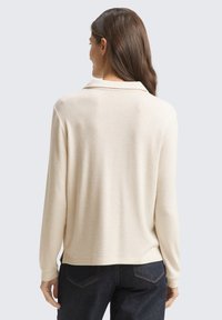 Beige Langarmpullover mit Kragen, aus weichem Stoff. Verfügt über eine lockere Passform und einen leicht abgerundeten Saum, kombiniert mit dunklen Jeans.