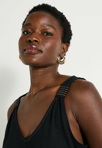 Top negro sin mangas con diseño de escote en V, que presenta detalles en hilo dorado en las tirantes. Textura suave y silueta ajustada.