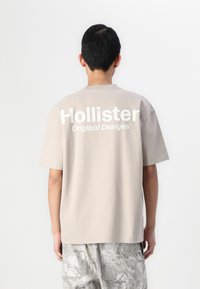 Beige Baumwoll-T-Shirt mit kurzen Ärmeln, auf dem großen weißen "Hollister"-Schriftzug und "Original Designs" auf der Rückseite zu sehen ist. Lockere Passform.