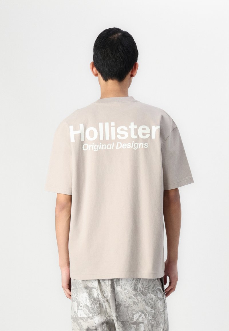 Beige Baumwoll-T-Shirt mit kurzen Ärmeln, auf dem großen weißen "Hollister"-Schriftzug und "Original Designs" auf der Rückseite zu sehen ist. Lockere Passform.