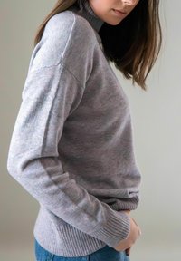 Vrouw in een lichtgrijze gebreide trui met geribbelde manchetten en zoom, staande in profiel met haar handen in de zakken van blauwe jeans.