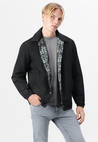 Veste noire avec un design à col et des poignets élastiques, dotée d'une doublure intérieure à motifs avec des motifs géométriques et floraux colorés.