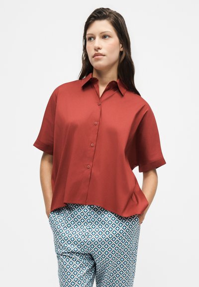 Femme debout, les mains dans les poches, vêtue d'une chemise rouge ample à manches courtes boutonnée et d'un pantalon bleu à motifs sur un fond uni.