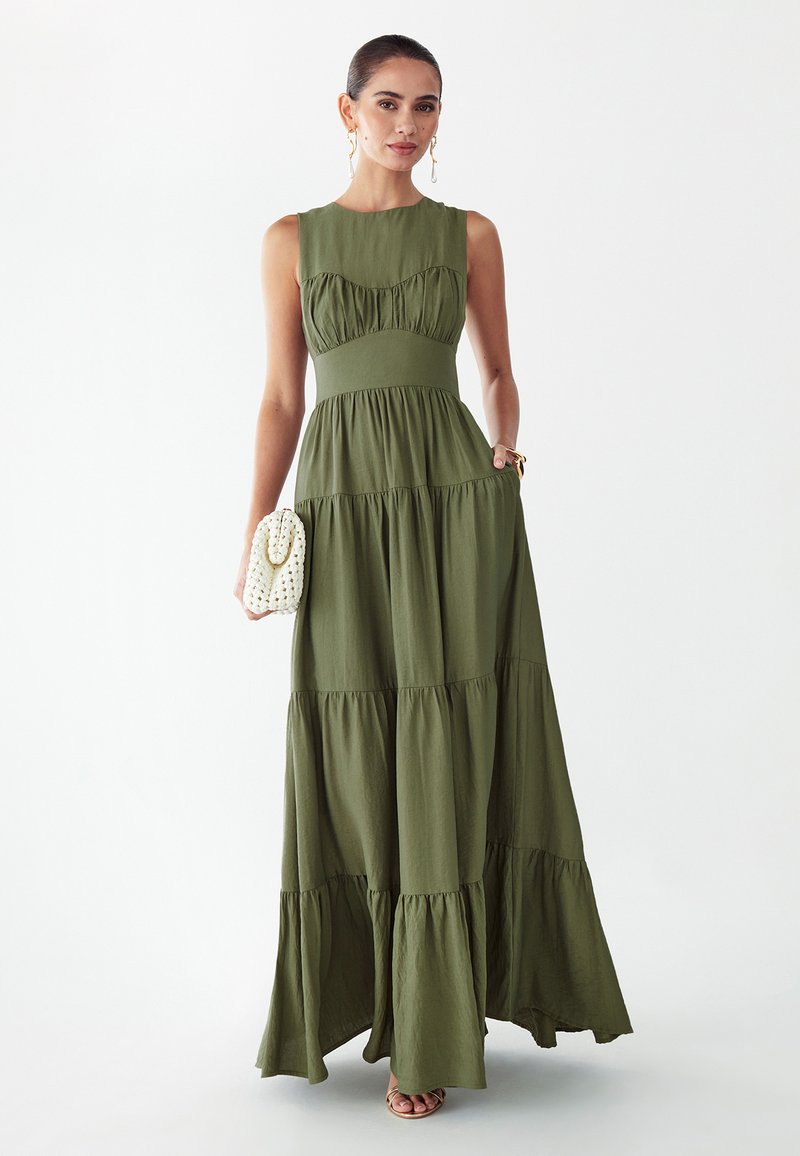 Vrouw draagt mouwloze olijfgroene gelaagde maxi-jurk, houdt witte geweven clutch vast, geaccentueerd met gouden oorbellen en armband.