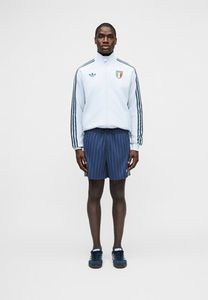 Άνδρας όρθιος, φοράει λευκό μπουφάν με φερμουάρ, μπλε ρίγες και έμβλημα, σκούρο μπλε σορτς με ρίγες και μπλε αθλητικά παπούτσια, σε λευκό φόντο.