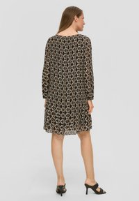 Zwarte doorschijnende jurk met een geometrisch patroon in beige en blauw. Heeft lange mouwen, een ronde halslijn en een losse pasvorm. Gecombineerd met hoge sandalen.