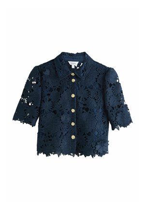 Femme aux longs cheveux blonds portant une robe midi en dentelle florale bleu marine et des sandales à talons noires, marchant devant un fond clair uni.
